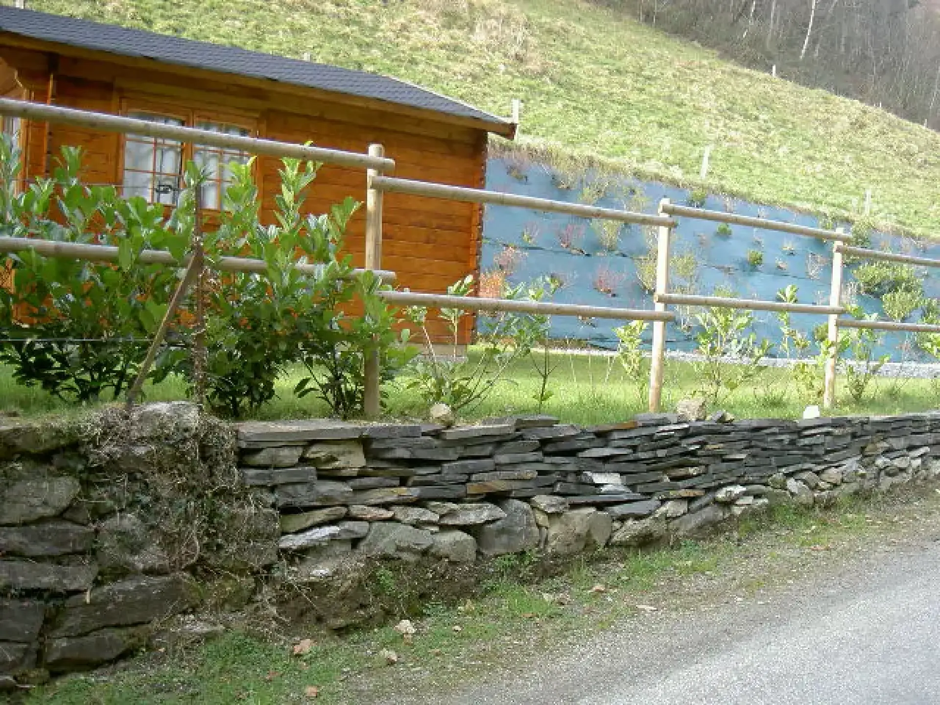 Aménagement extérieur avec muret en pierres et clôture en bois autour d’un chalet bordant un plan d’eau.