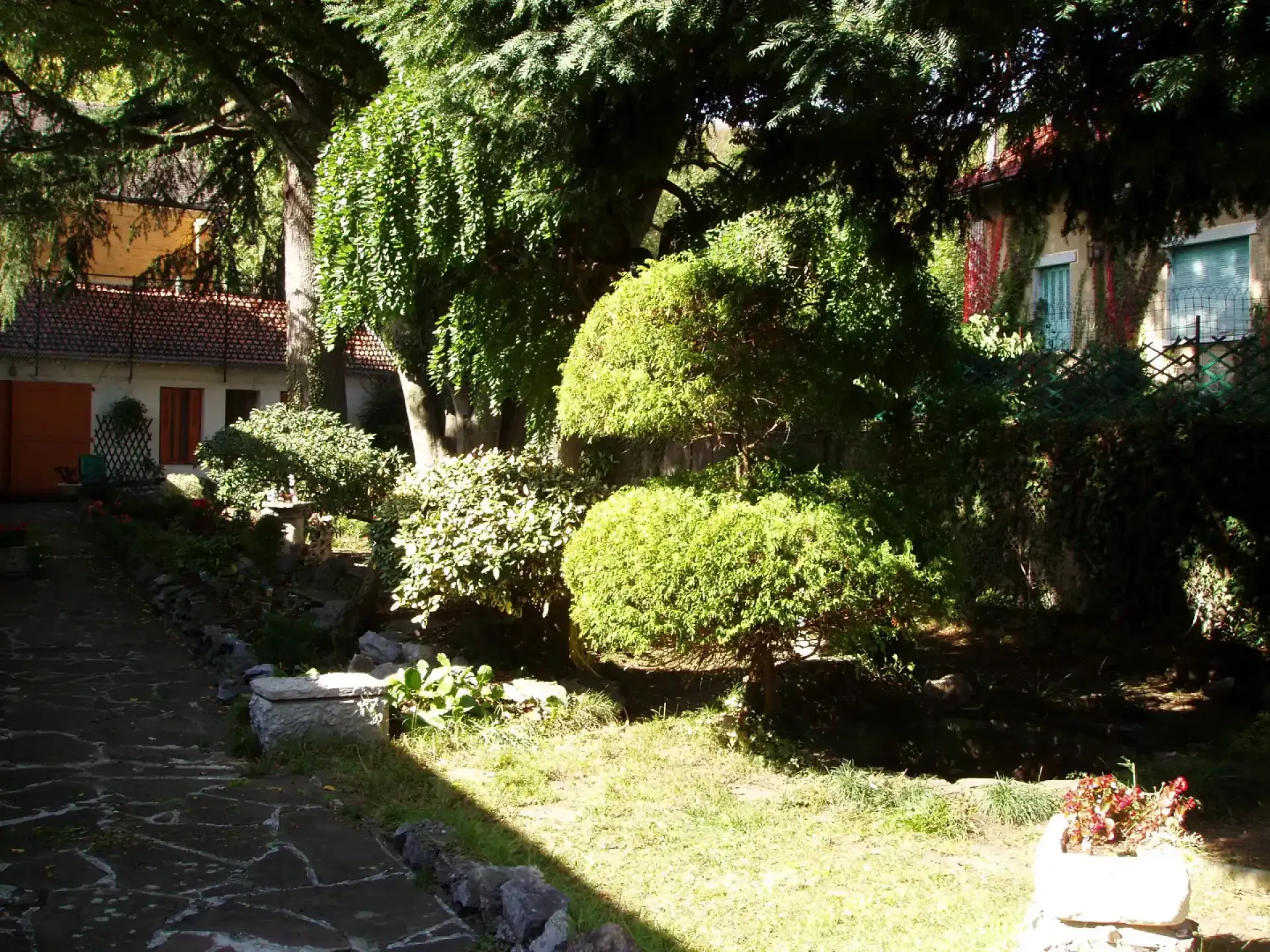 Aménagement paysager d'un jardin arboré avec arbustes taillés, massifs fleuris et chemin en pierre.